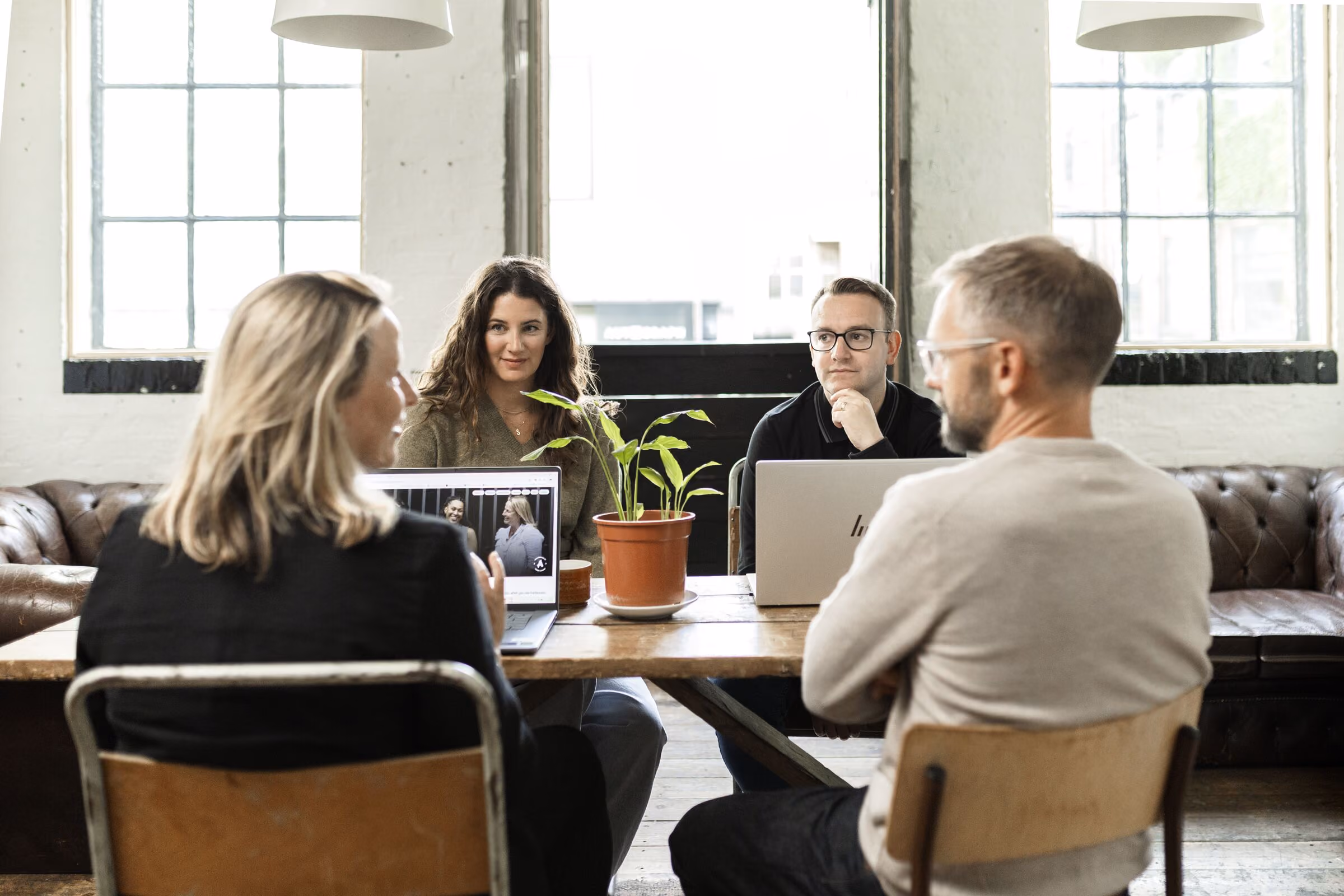Four people sitting around a wooden table in a modern, light-filled office. Two face the camera, one woman looks at a laptop screen showing a photo of two people, and a man with glasses and a beard is seated with folded arms. A potted plant is in the center of the table.