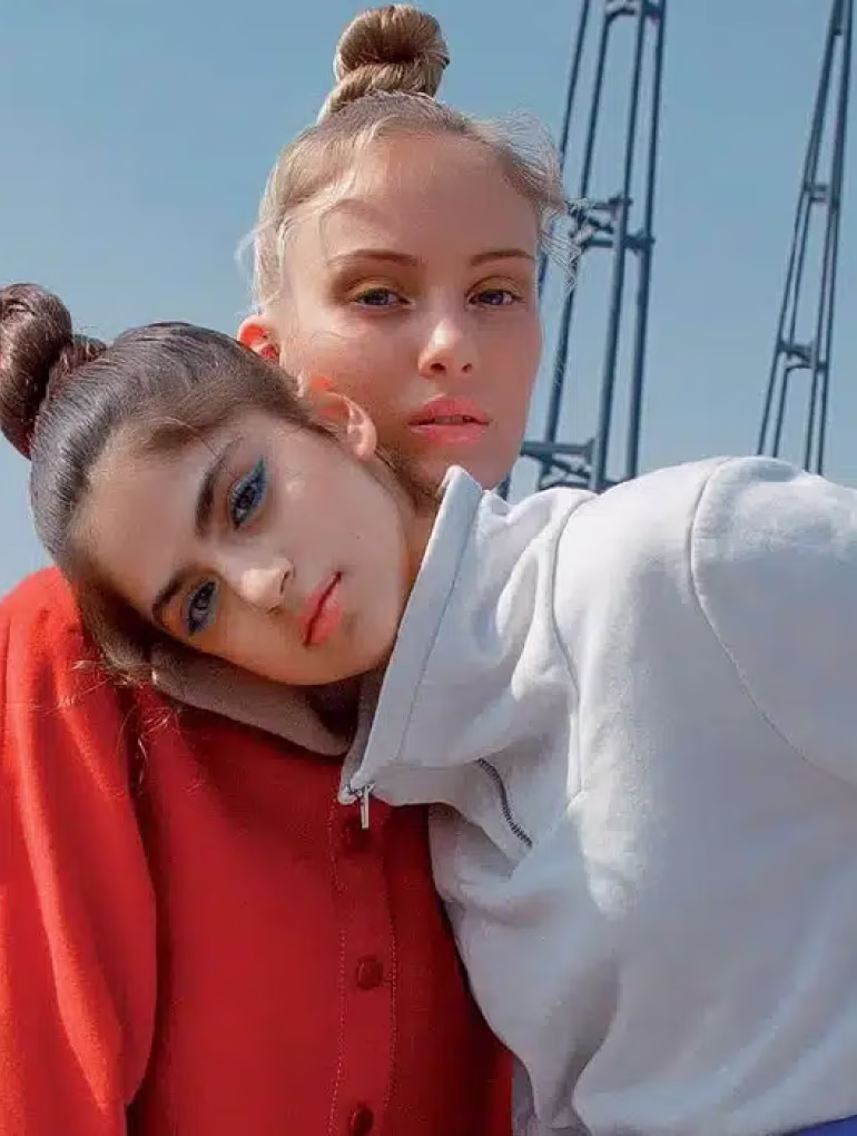 Two young women with buns, one resting head on the other's shoulder, wearing red and white tops outdoors.