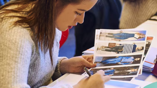 Woman examining fashion design sheets featuring models in various outfits while writing.