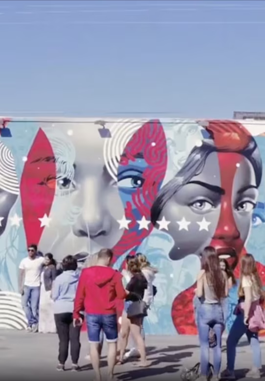 People walking and standing in front of a colorful mural featuring stylized faces with stars.