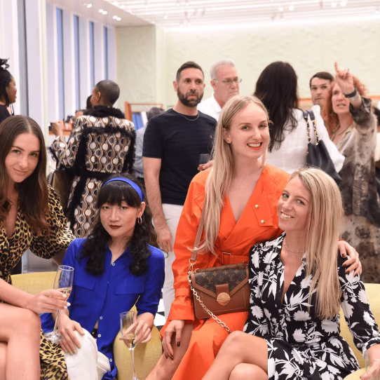 Group of people socializing indoors, three women sitting in the front holding drinks, smiling.