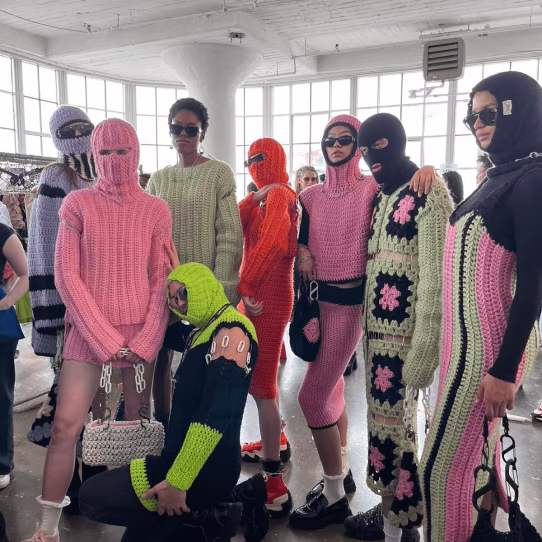 Group of people wearing colorful knitted outfits and balaclavas in a bright room with large windows.