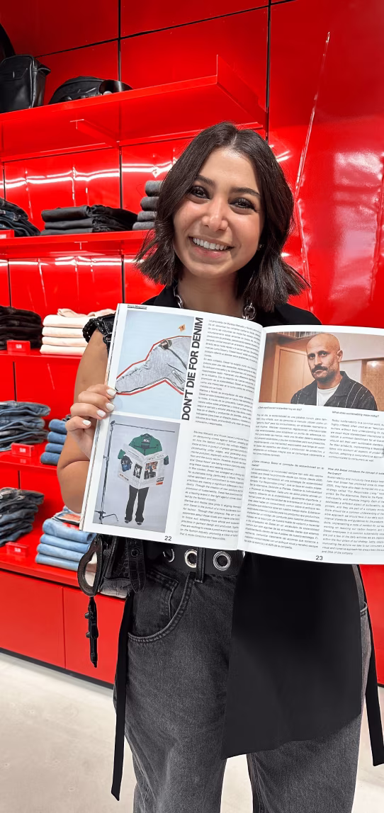 Smiling woman holding open magazine with text and photos in a store with red shelves and folded clothes.