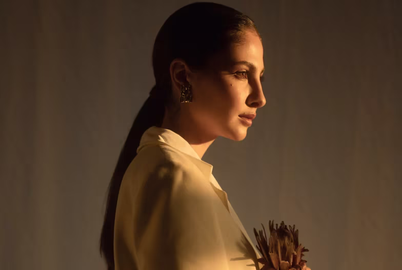 Profile of a woman with slicked-back hair wearing earrings and a beige jacket in soft warm lighting.