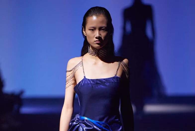 Woman in dark blue satin dress with chain shoulder details walking on dimly lit runway.