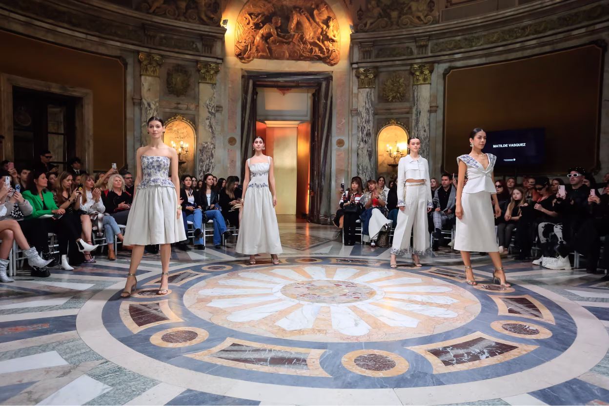 Four fashion models in white and silver outfits standing in a grand hall with marble floor and seated audience.