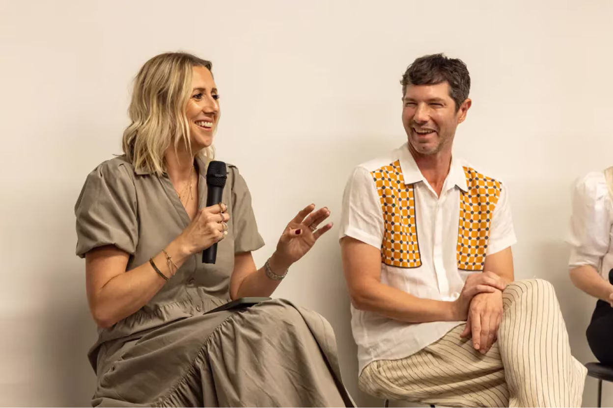 Smiling woman holding a microphone talks to a man in a white shirt with orange patterned patches.