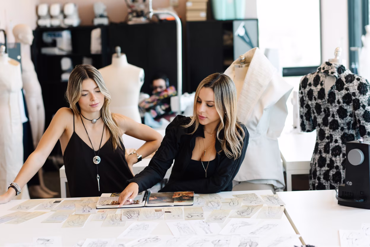 Two women reviewing fashion sketches and designs at a large white table in a design studio.