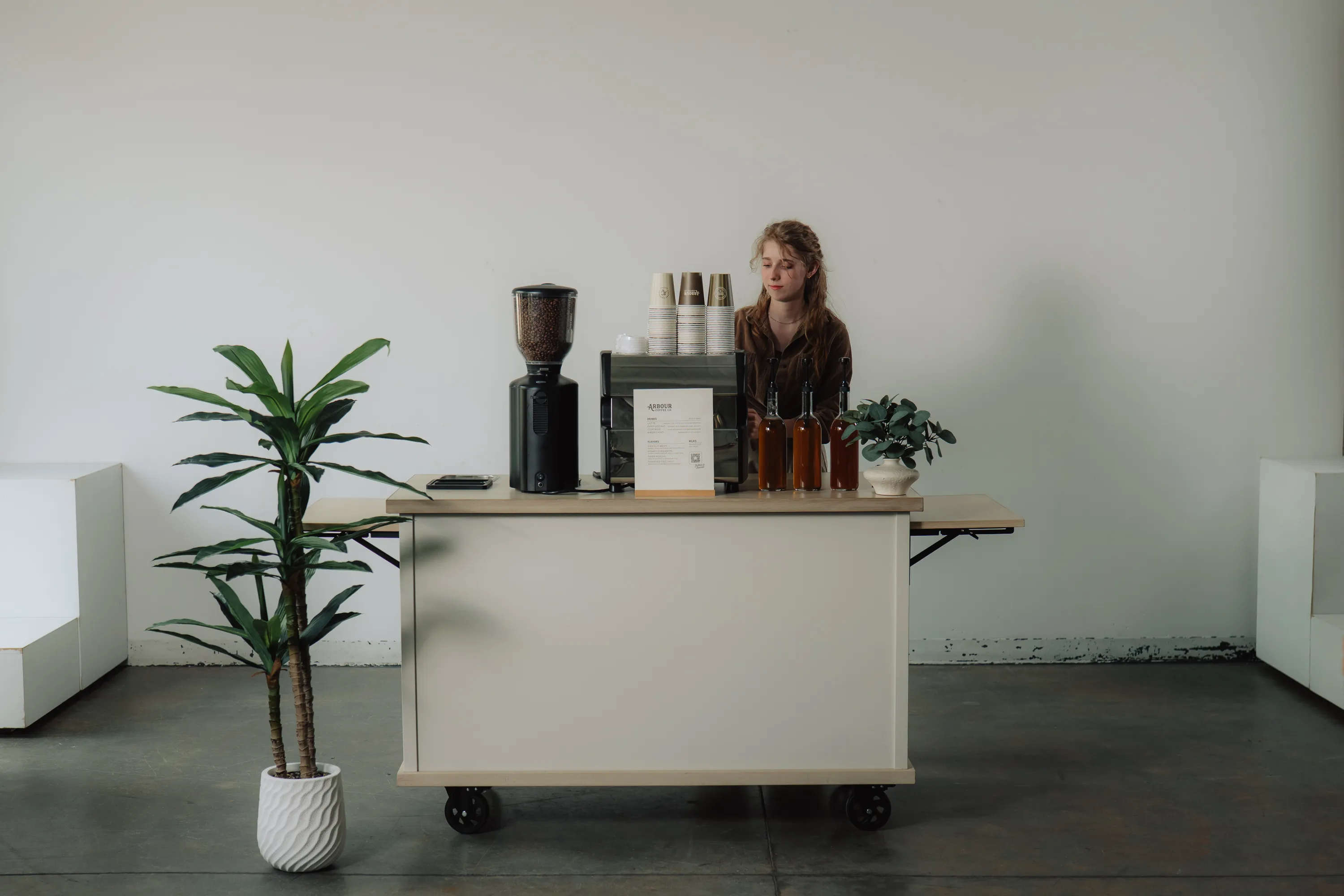 A barista stands behind a sleek white mobile coffee cart with espresso maker and coffee grinder.