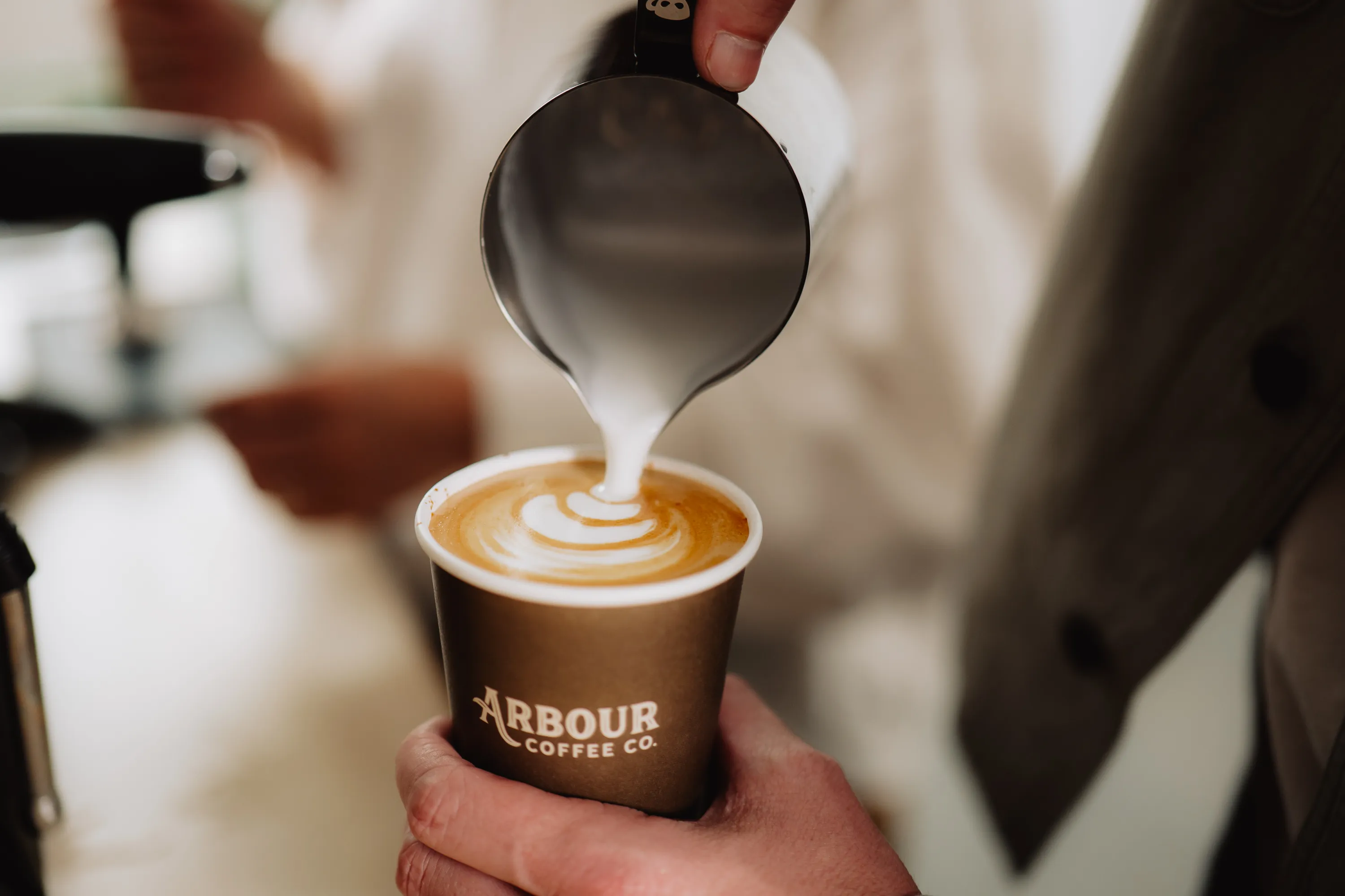 A barista pouring steamed milk into an Arbour Coffee Co. paper cup to create latte art.