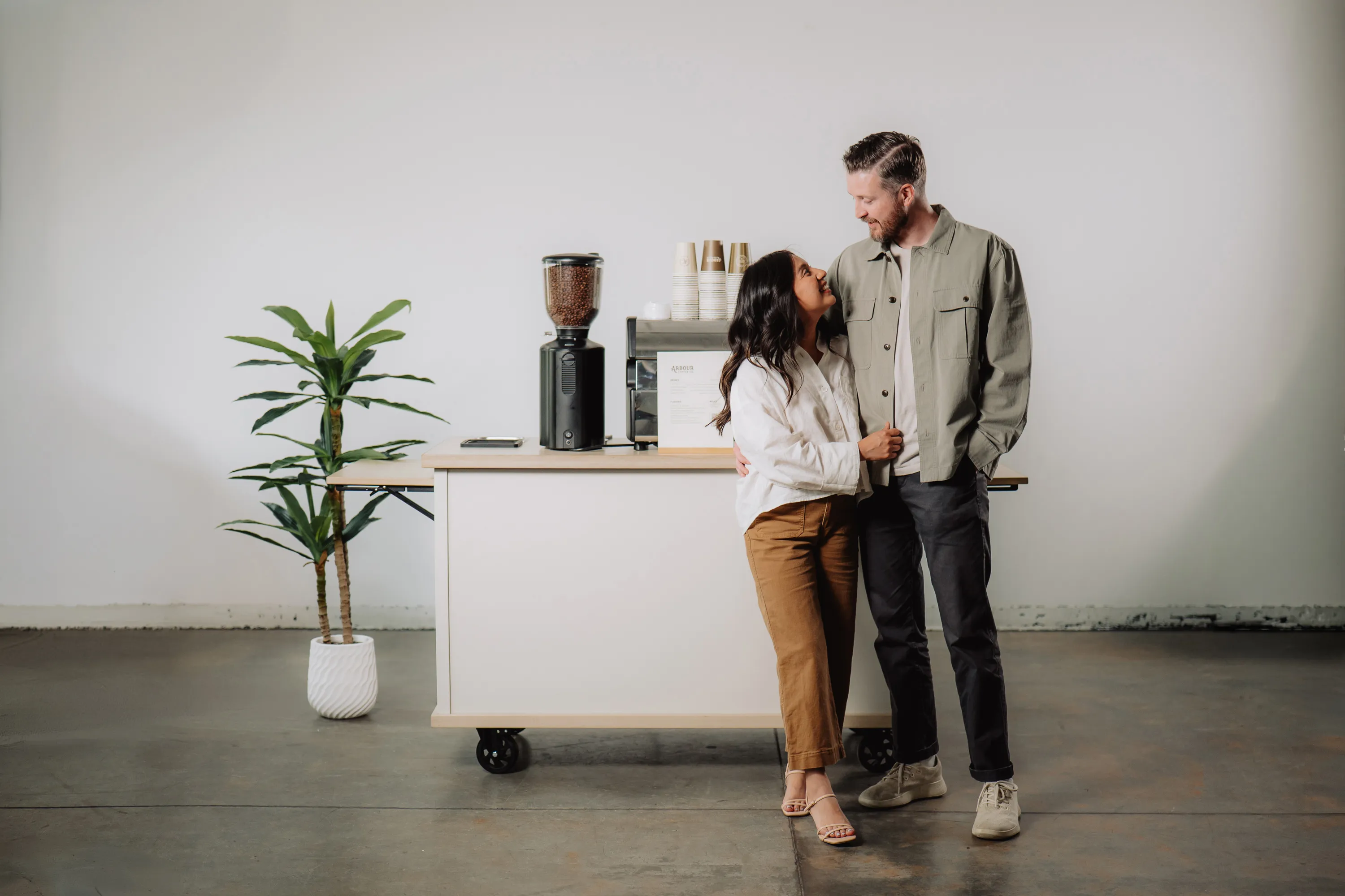 A smiling couple posing in front of a mobile espresso coffee cart in a minimalist white studio.