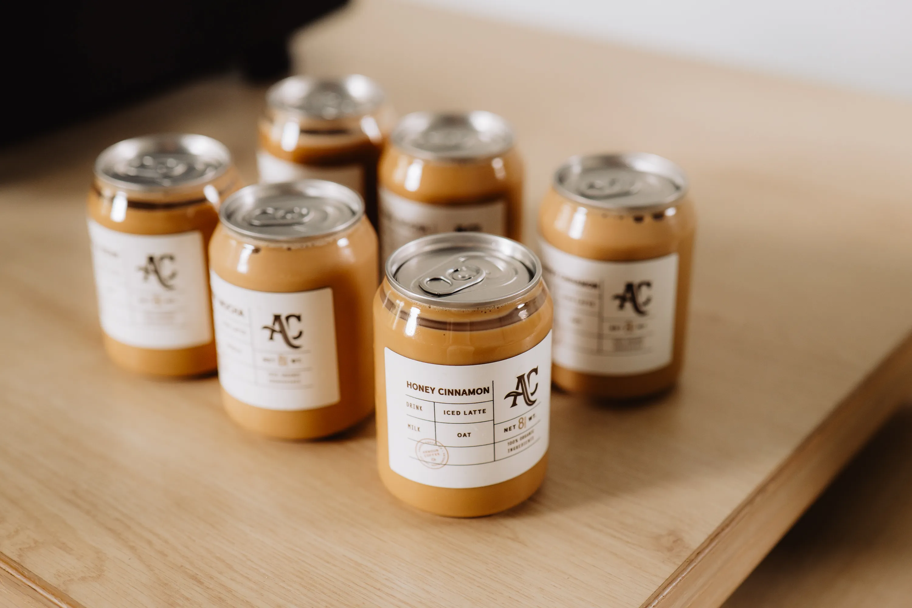 Six canned Honey Cinnamon iced latte drinks with oat milk on a light wood surface.