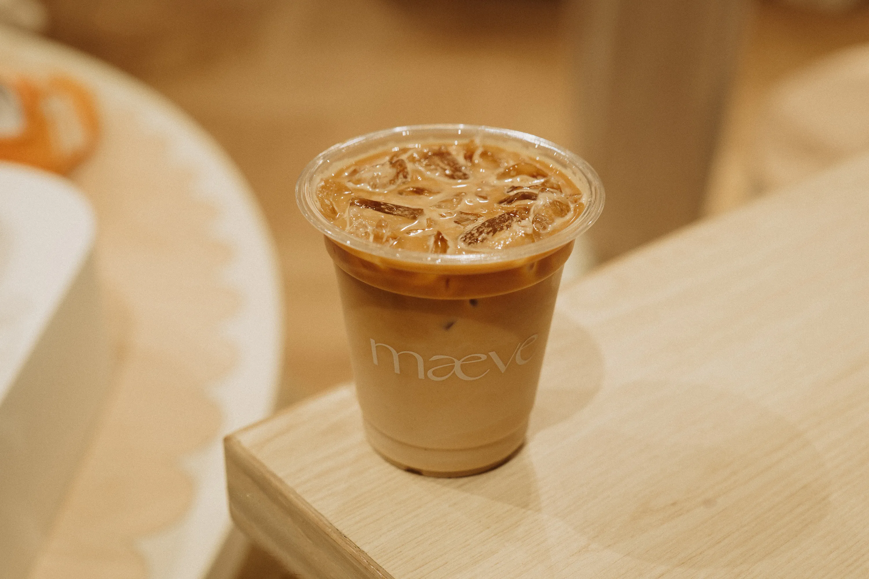 An iced latte in a clear plastic cup branded with the Maeve logo on a light wood table.