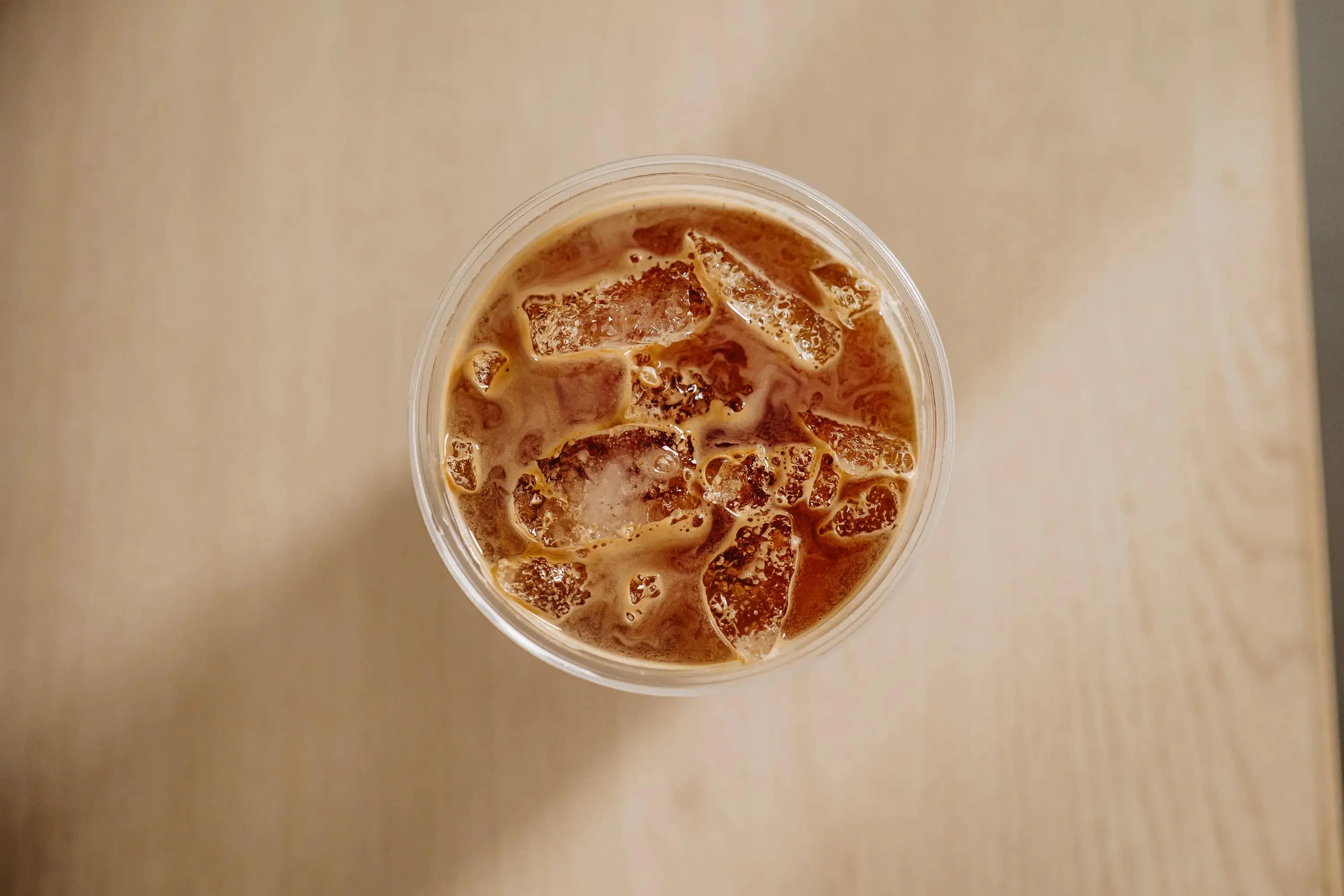 Top-down view of iced coffee with ice cubes in a clear plastic cup on a light wooden table.