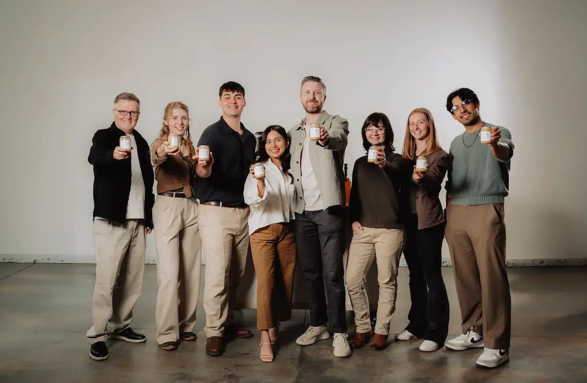 A group of eight smiling people in a studio posing together and holding up small jars of product.