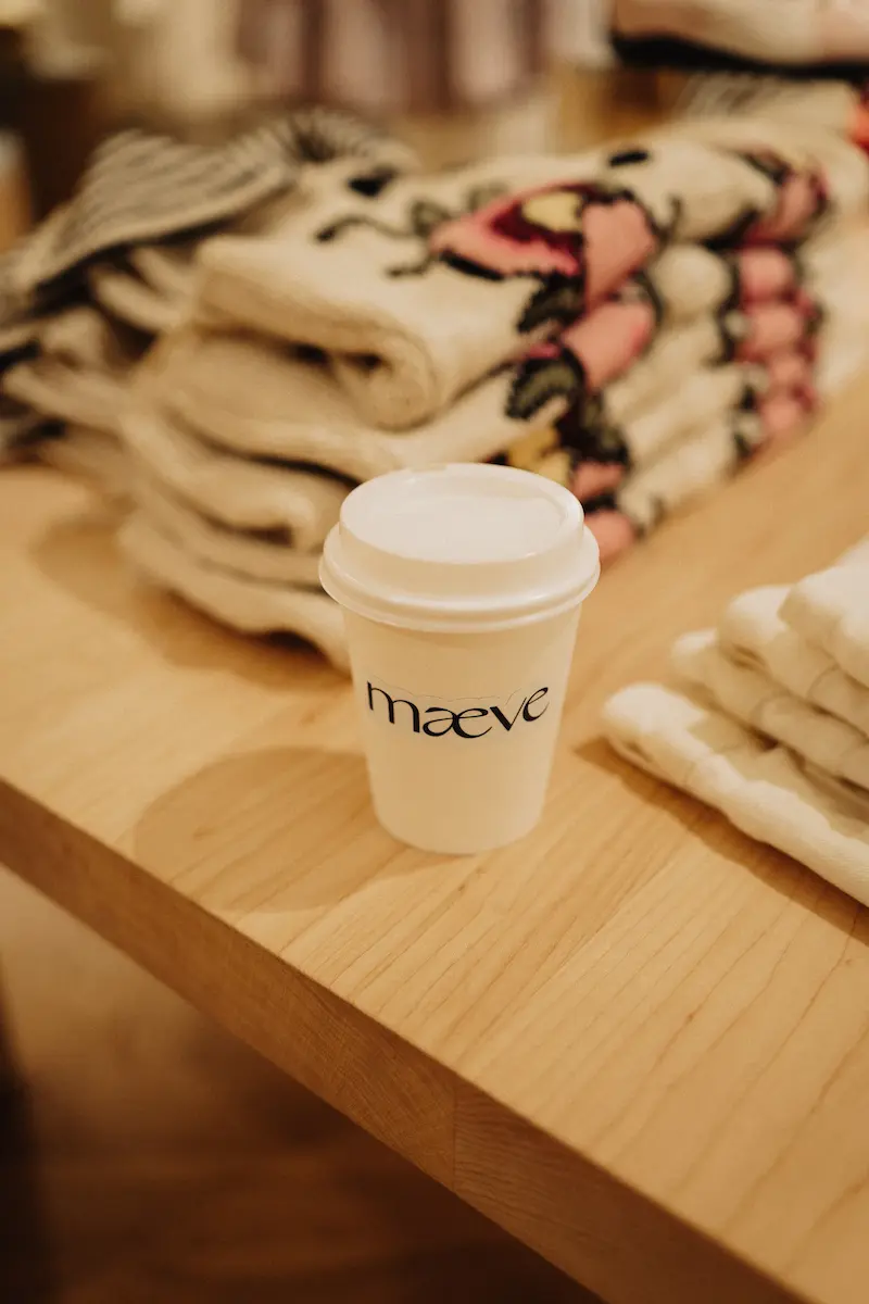 A white paper coffee cup with Maeve branding sitting on a wooden table next to stacked folded sweaters.