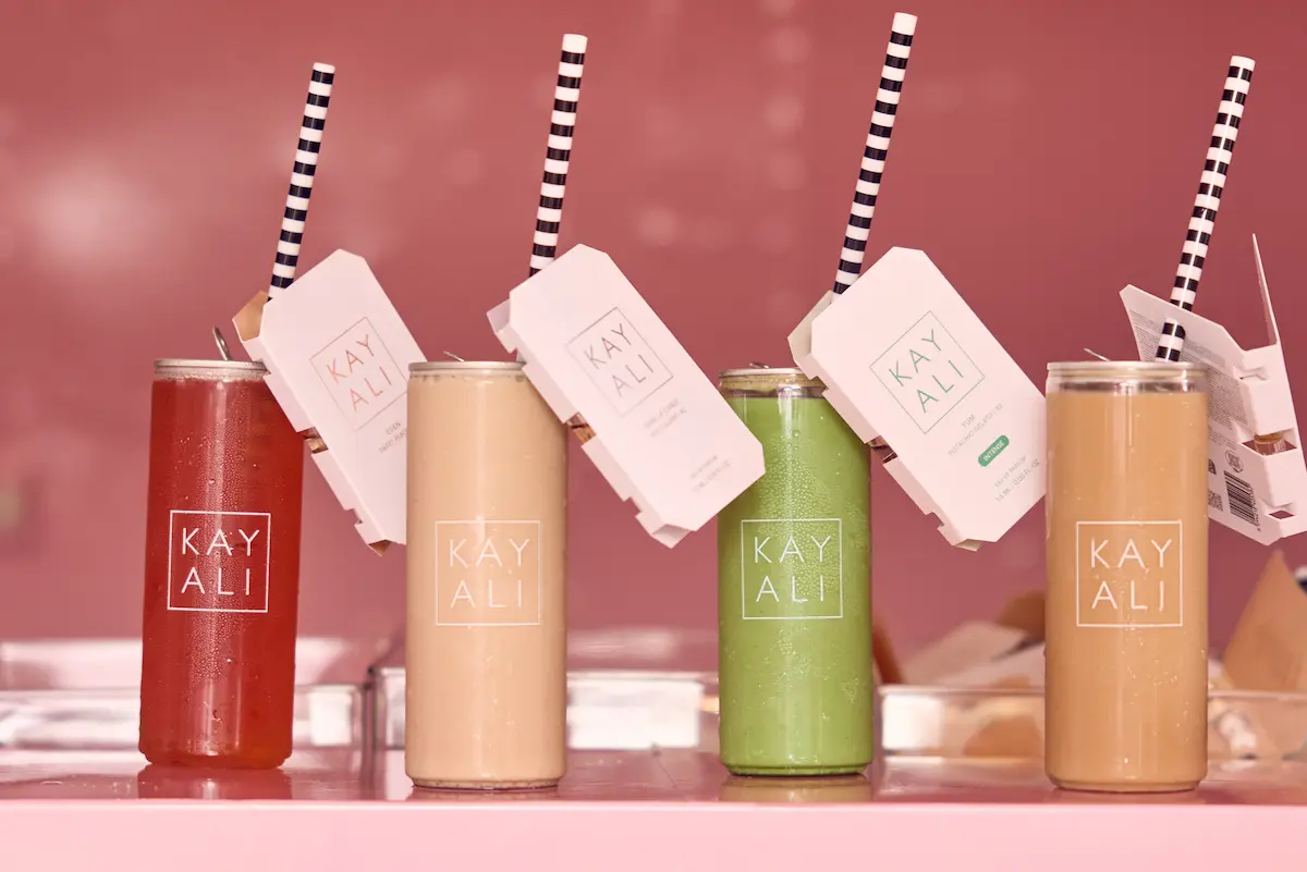 Four Kayali-branded canned drinks in red, cream, green and caramel, each with a scent card on the straw