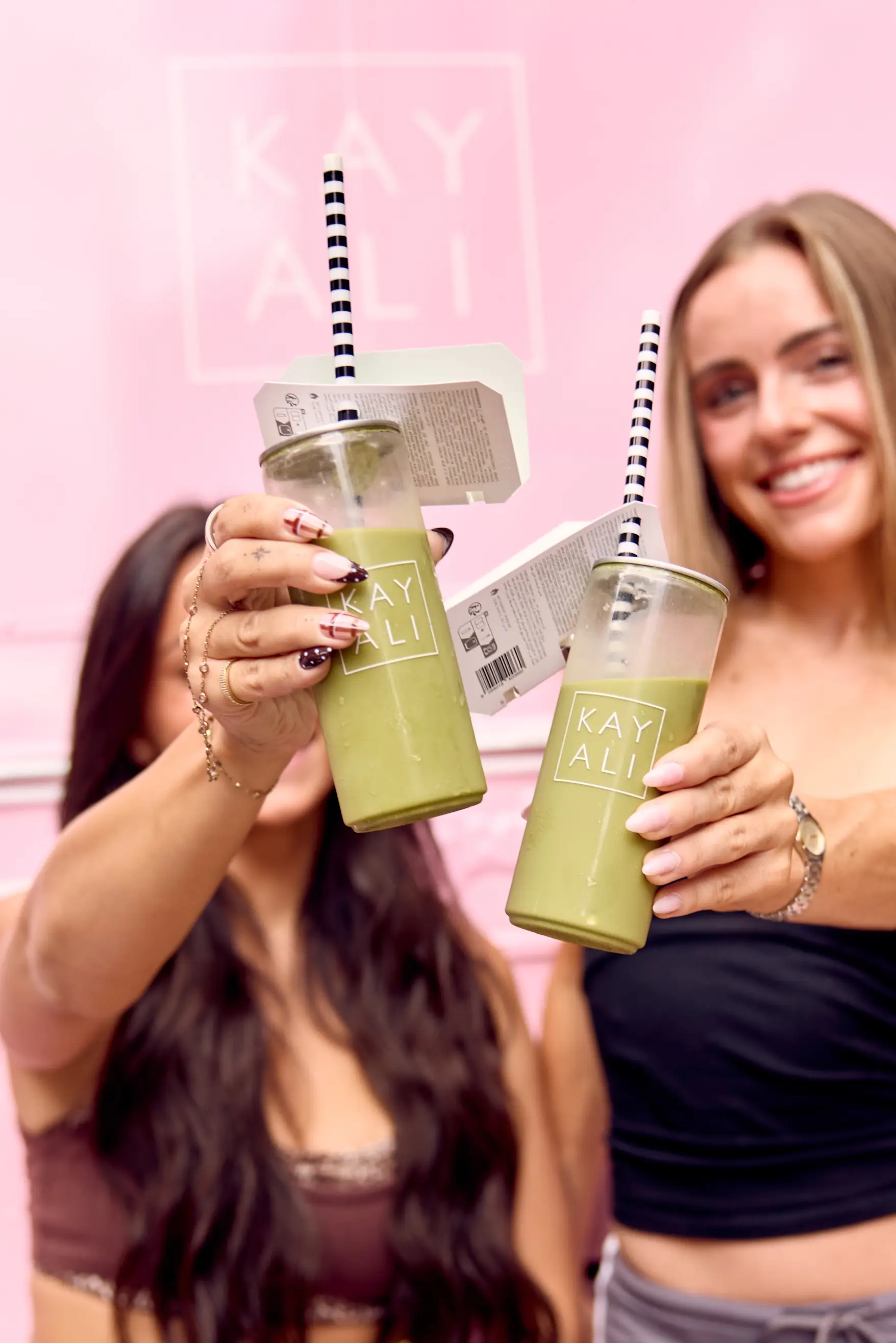 Two guests raising Kayali-branded canned matcha lattes at a pink brand pop-up.