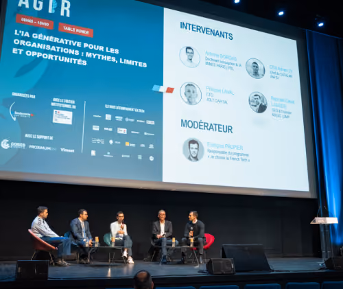 Cinco hommes assis sur scène discutant lors d'une table ronde devant un écran affichant les intervenants et le modérateur.
