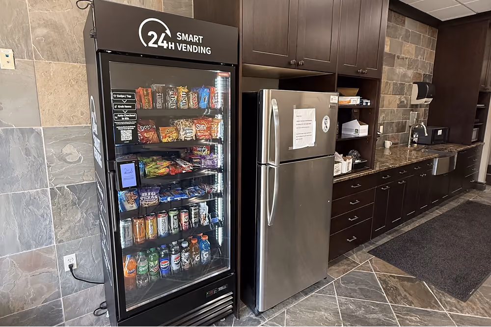A 24-hour smart vending machine stocked with snacks and drinks next to a stainless steel refrigerator in a kitchen.