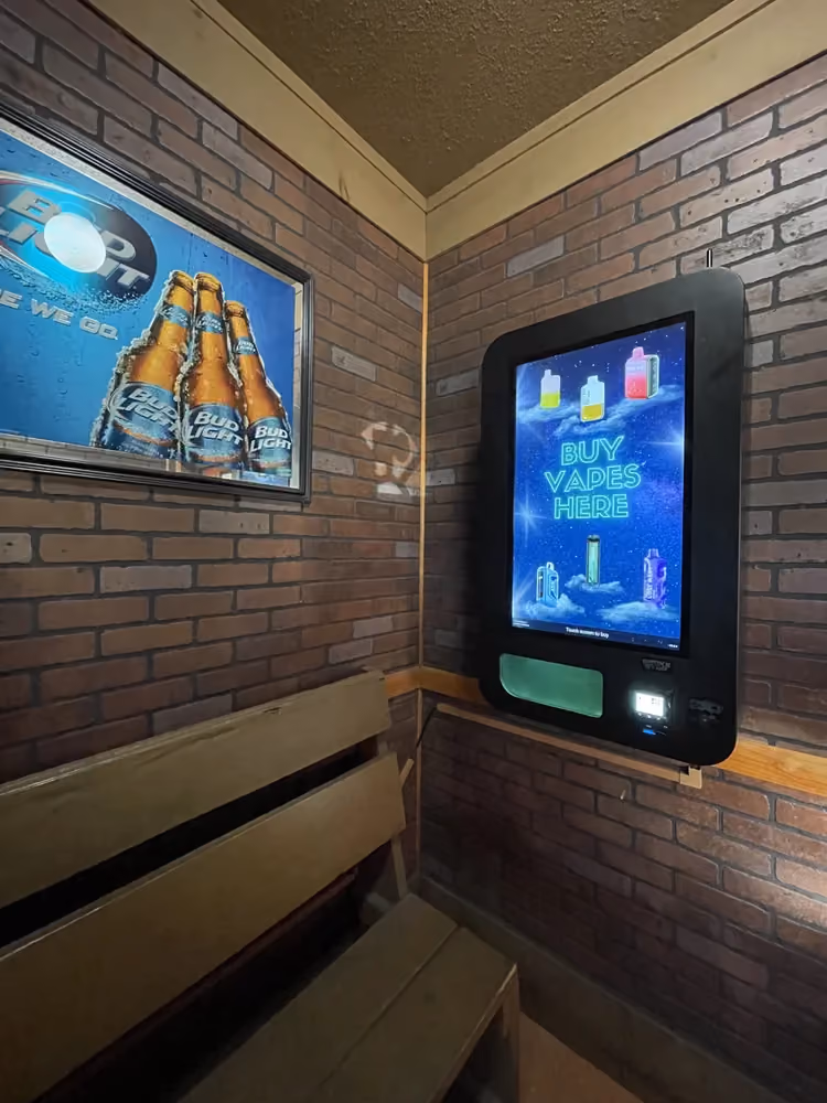 Small corner room with brick walls, wooden bench, Bud Light ad, and a vape vending machine with 'Buy Vapes Here'.