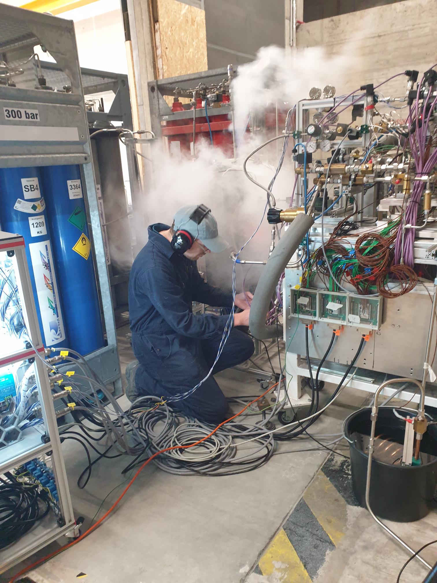 Technician in a cap and ear protection kneeling among cables, working on industrial equipment with steam nearby.