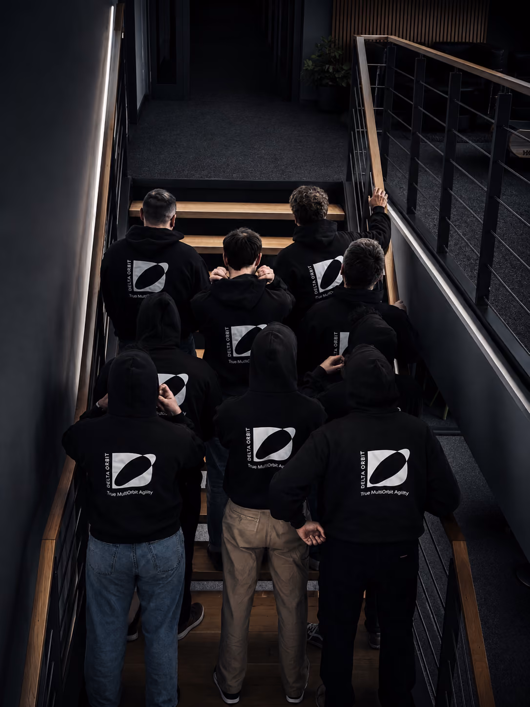 Seven people in black hoodies with Delta Orbit logos standing on stairs facing away.