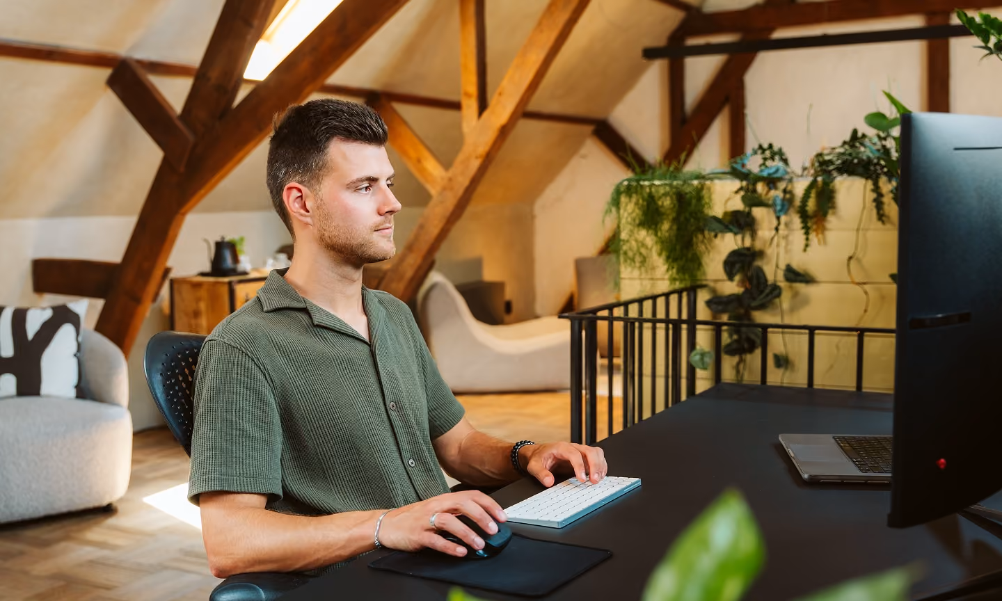 Man in green shirt sitting at desk using computer mouse and keyboard in a cozy room with plants.