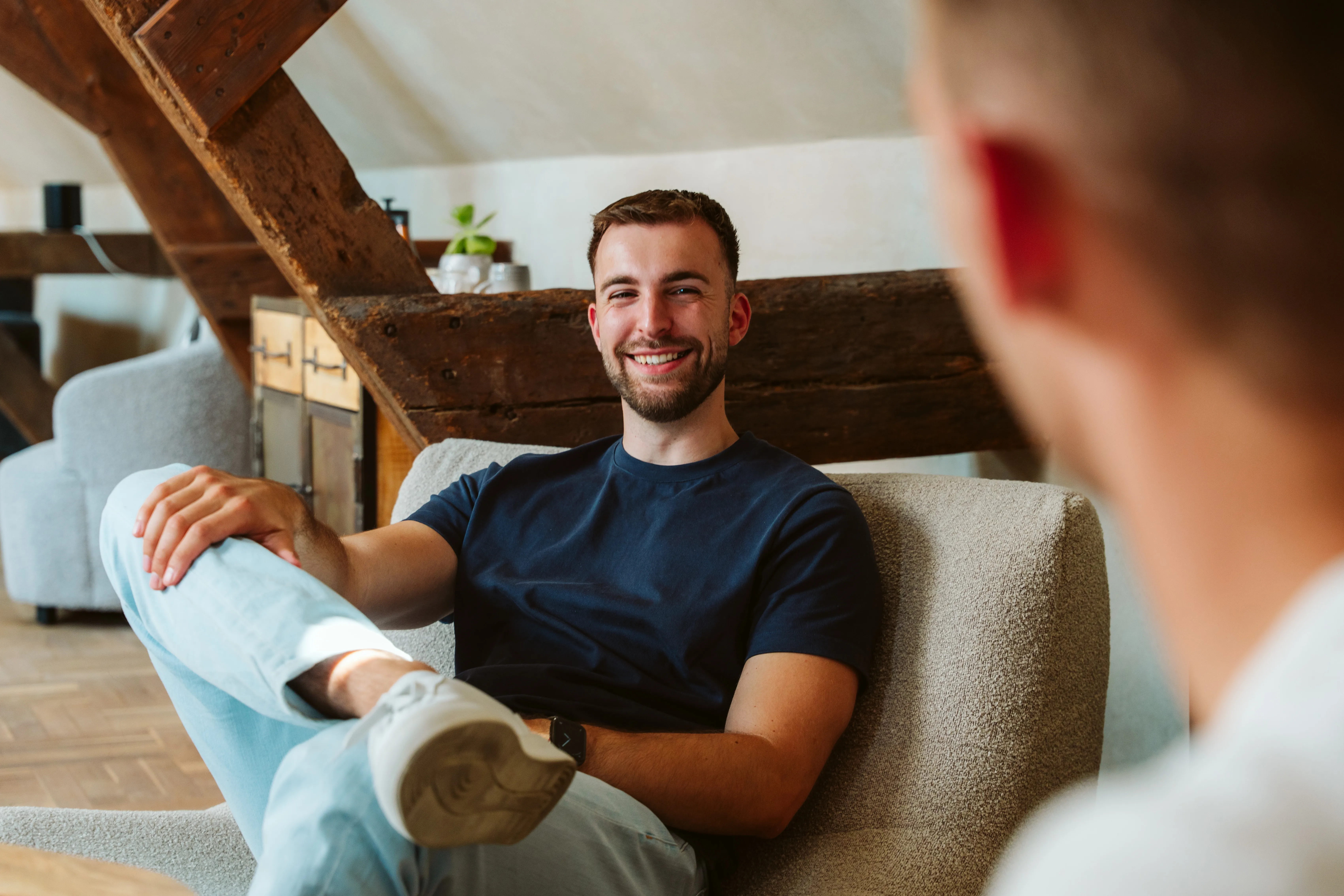 Man lacht zittend op een beige stoel met één been over de knie, in een warme, rustieke kamer.