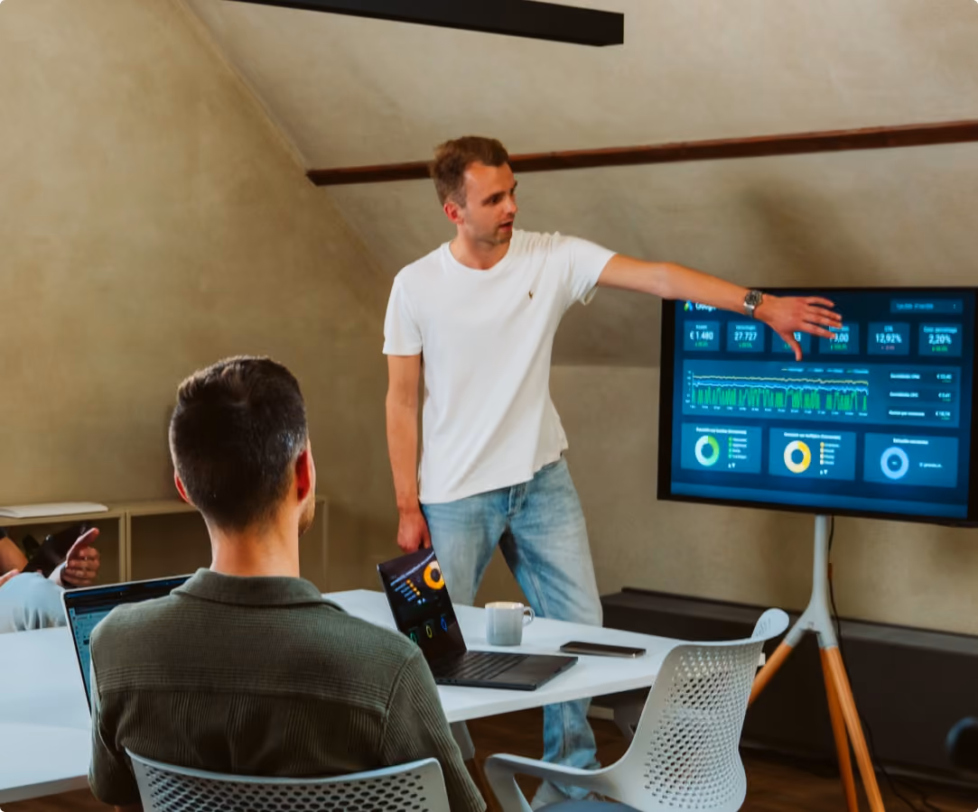 Man in wit t-shirt presenteert grafieken en statistieken aan groep in vergaderruimte met laptops.