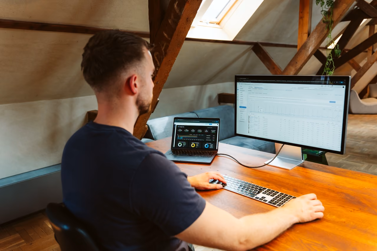 Man werkt thuis op een houten bureau met laptop en grote monitor met data-analyses.