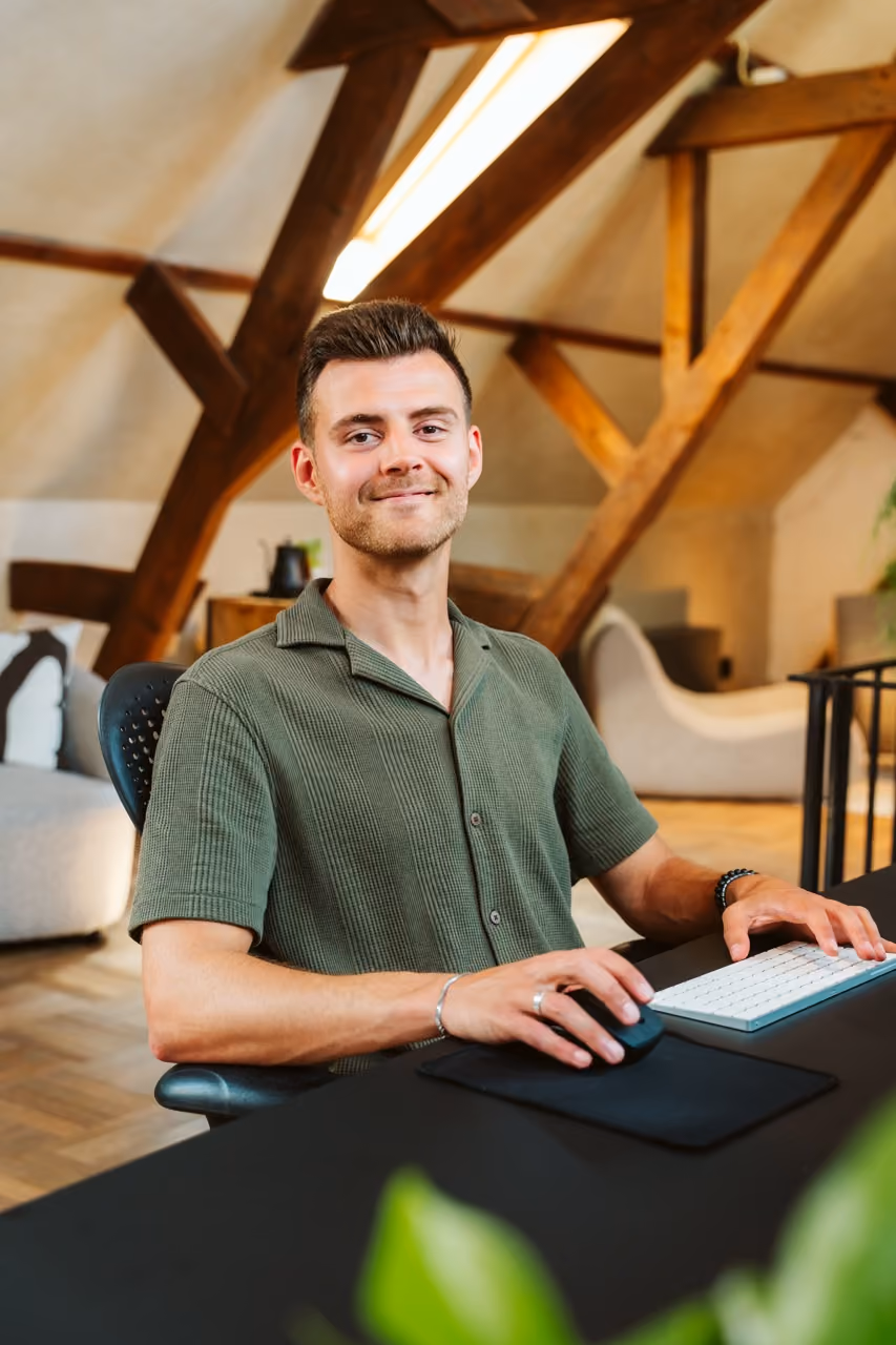 Jonge man zittend aan bureau met computer in een kamer met houten balken.