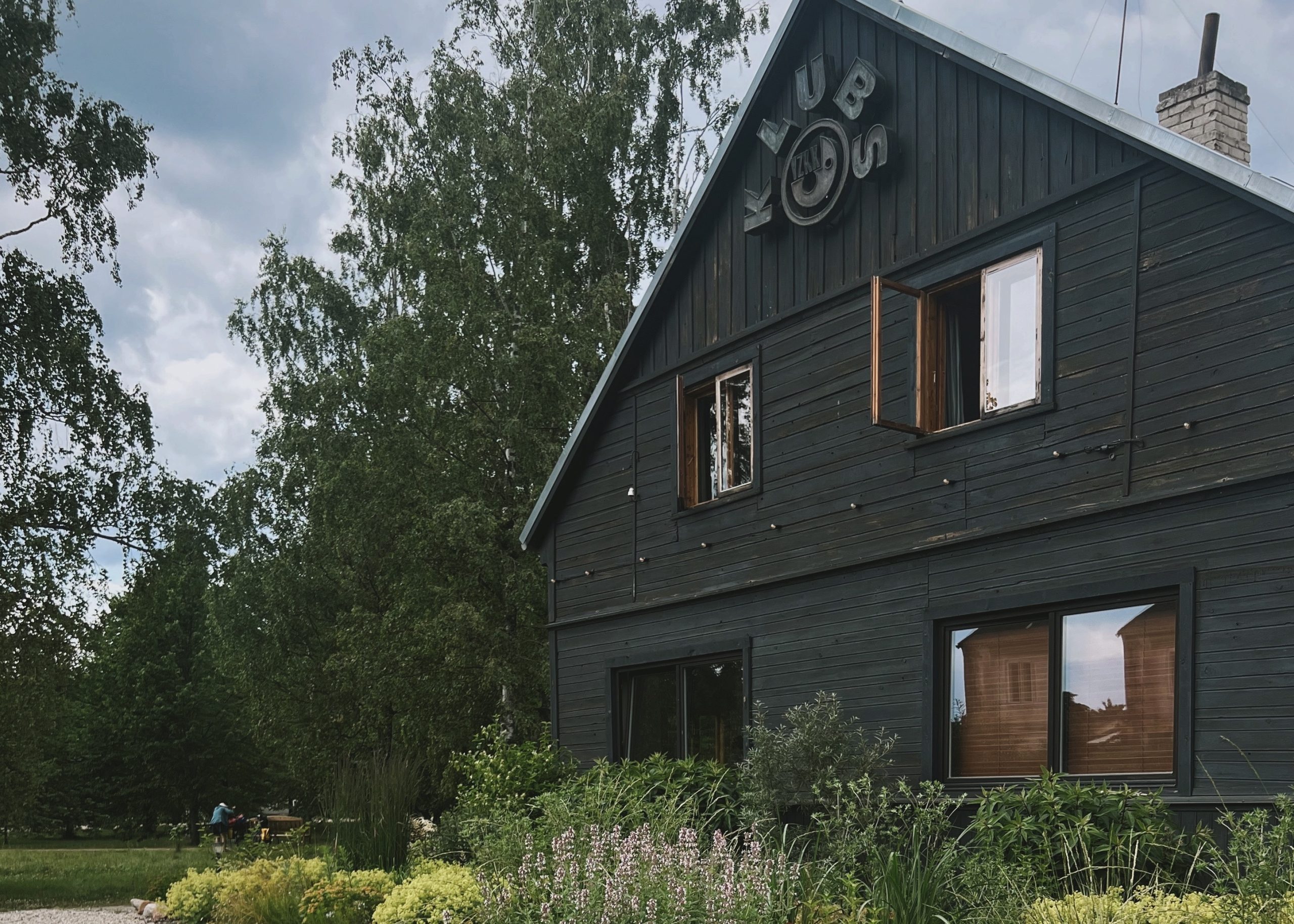 A dark wooden house with the word "KUBO" on the gable, set beside tall trees and a garden with grasses and shrubs.