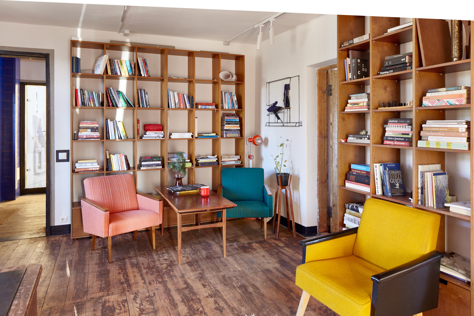 A cozy reading room with three colorful armchairs, wooden bookshelves filled with books, and a small coffee table on a dark wood floor.