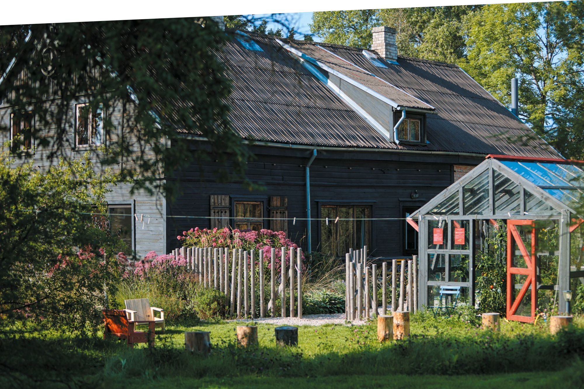 A dark wooden house with a sloped metal roof stands behind a fenced garden, with a small greenhouse in the foreground.