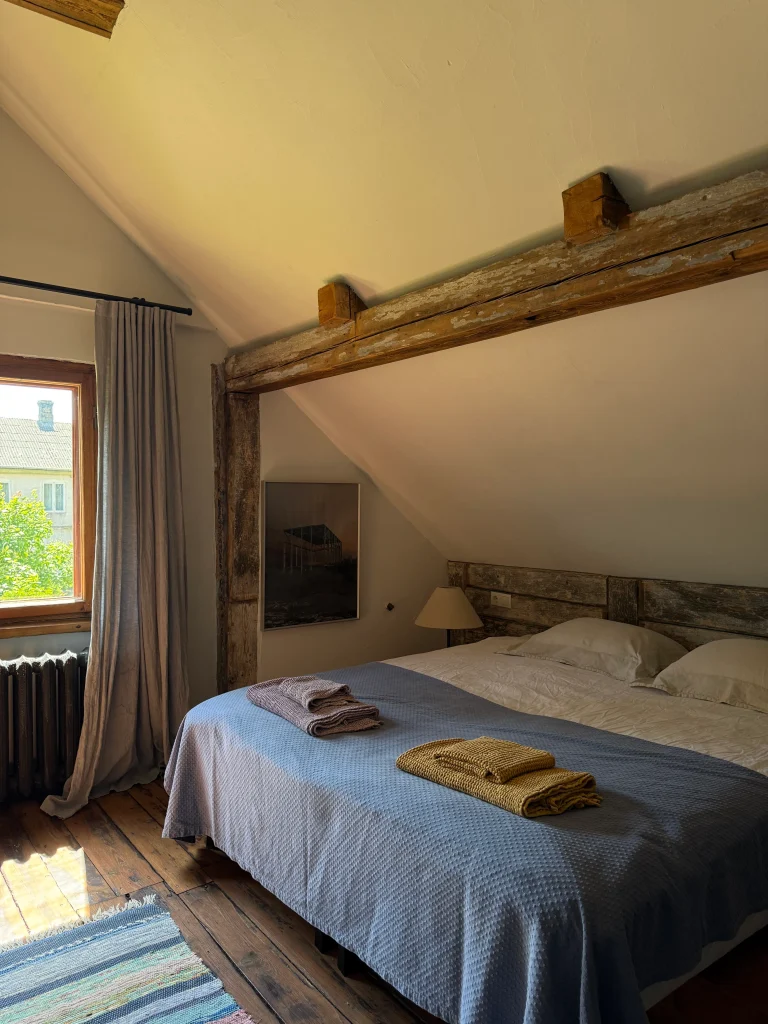 A rustic bedroom with a bed covered in a blue quilt and folded towels at the foot, beside a window with gray curtains and a sunlit wooden floor.
