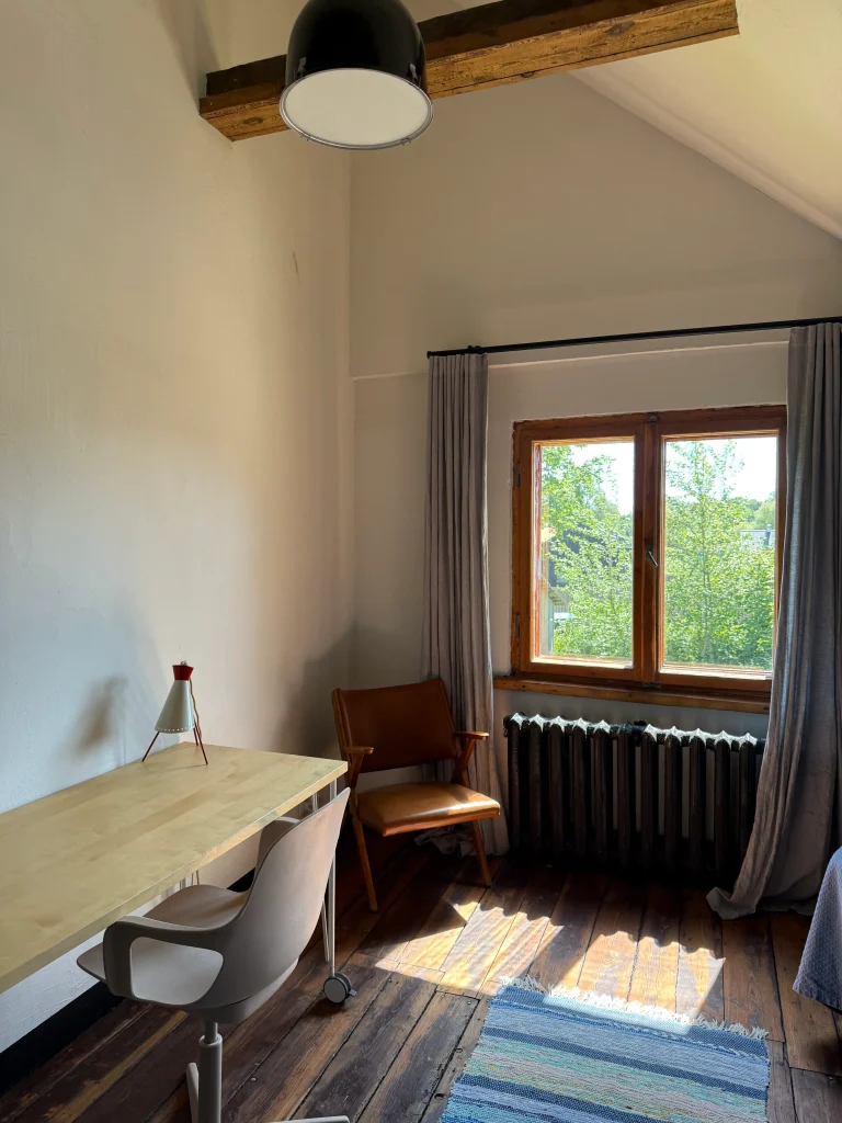 A small room with a wooden desk, chair, and lounge chair beside a window with gray curtains and a black radiator, with sunlight on the hardwood floor.