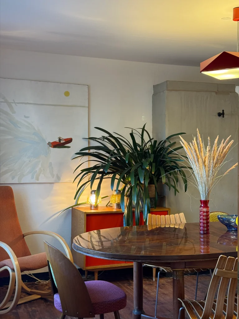 A round wooden dining table with mixed chairs sits in a cozy room, with a large potted plant, a red cabinet, and a tall vase of dried grasses behind it.