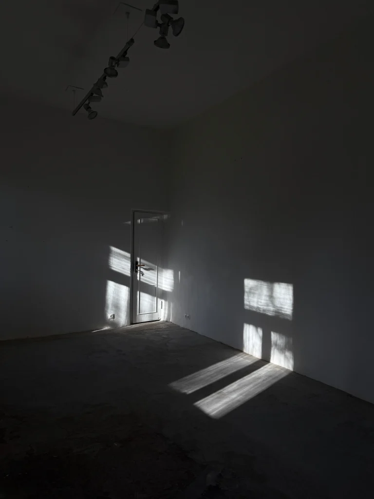 An empty dark room with white walls, a glass door, and sunlight casting rectangular shadows across the floor and wall.