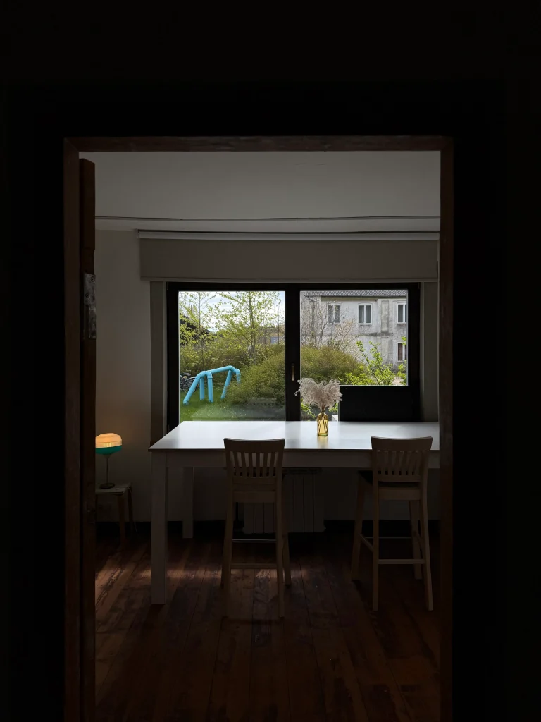 A dining table with two wooden chairs sits by a large window, with a small vase of flowers and a blue playground structure visible outside.