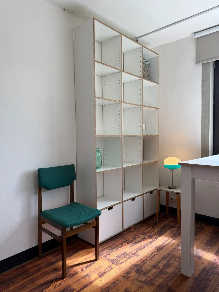 A tall white shelving unit with open compartments and lower cabinets stands beside a green upholstered chair, with a small side table, lamp, and desk in a bright room with wood flooring.