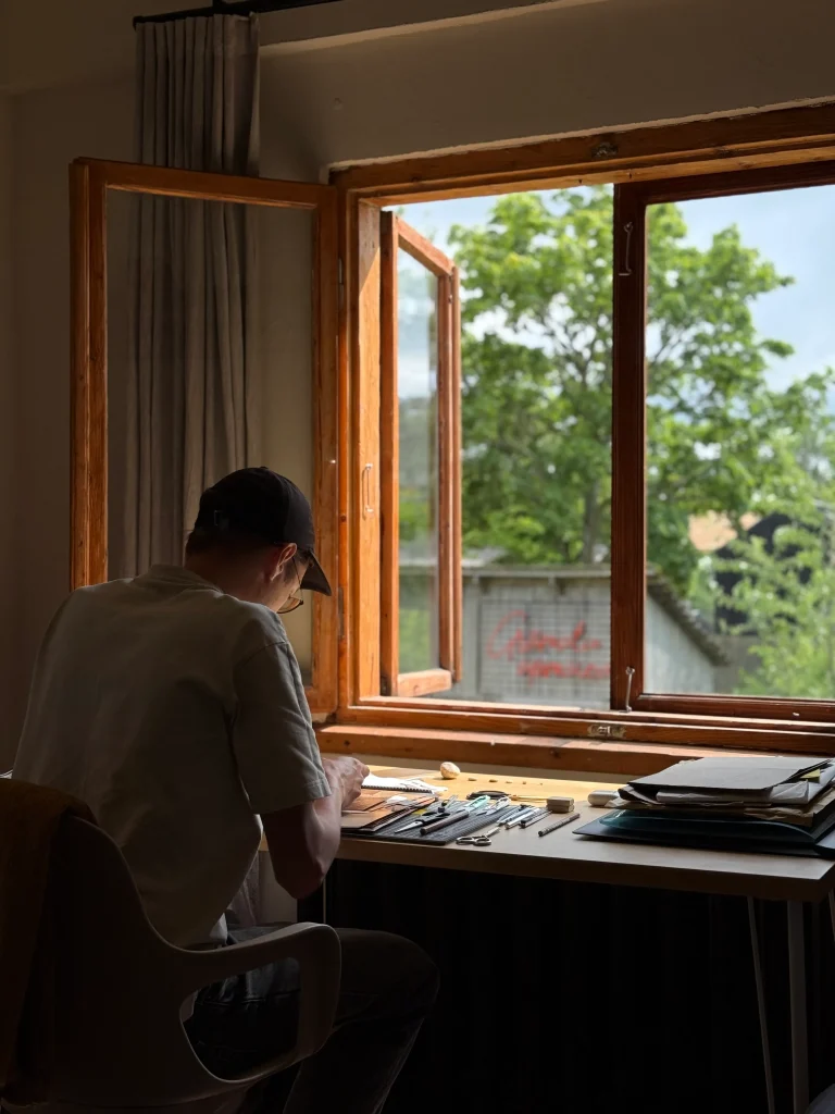 A man in a cap sits at a desk by an open wooden window, working amid papers and tools with green trees visible outside.