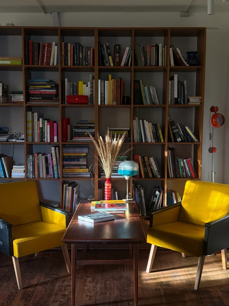 Two yellow armchairs face a wooden coffee table in front of a tall bookshelf filled with books, with a vase of dried grass and a blue glass lamp on the table.