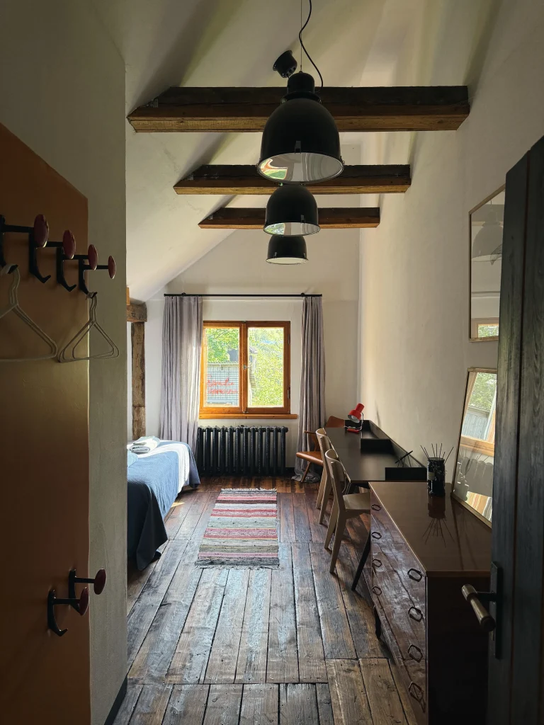 A narrow bedroom with a wooden floor and exposed ceiling beams, showing a bed on the left, a desk and chair on the right, and a window at the far end.