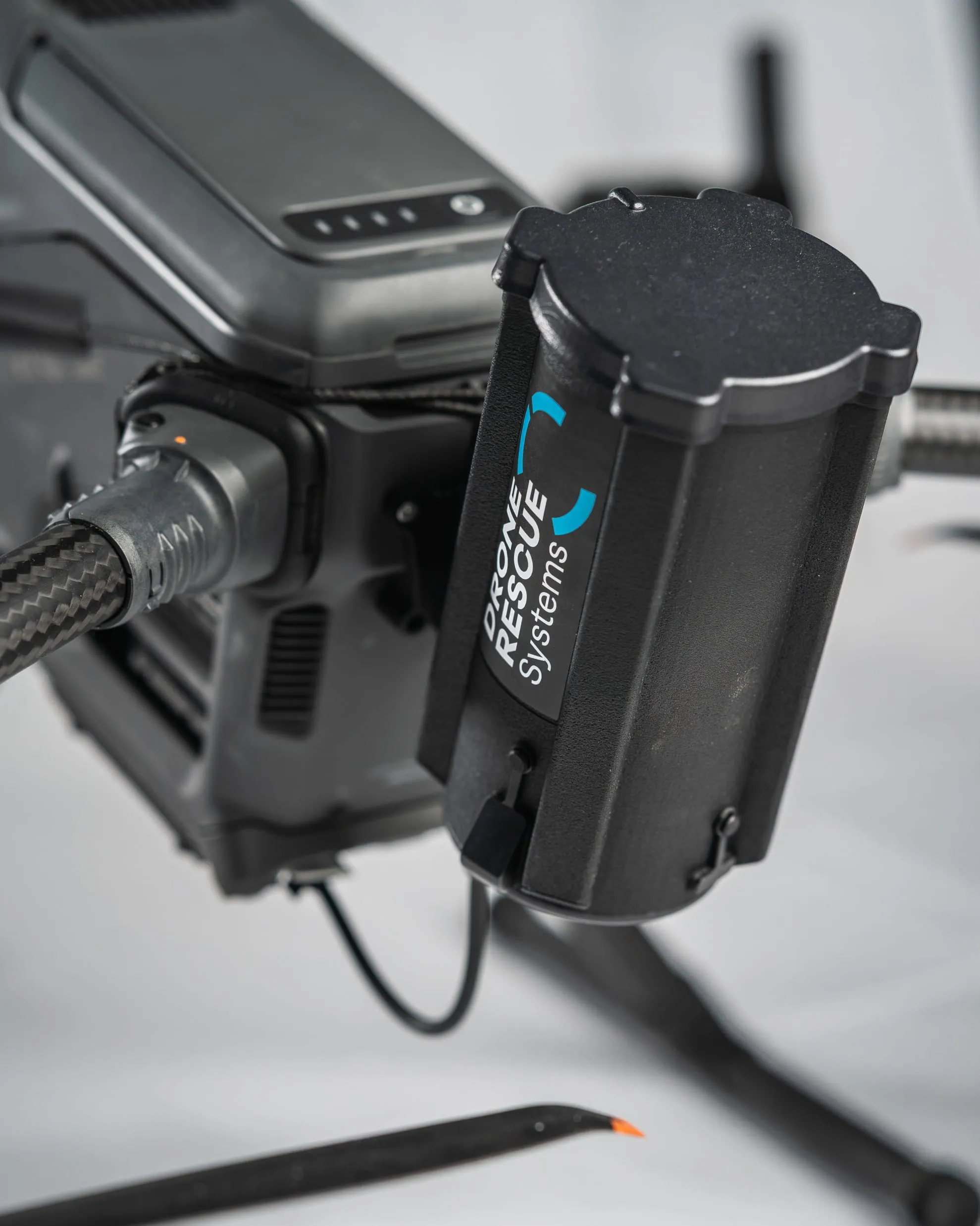 Close-up of a black Drone Rescue Systems device attached to a drone arm with carbon fiber pattern cable.