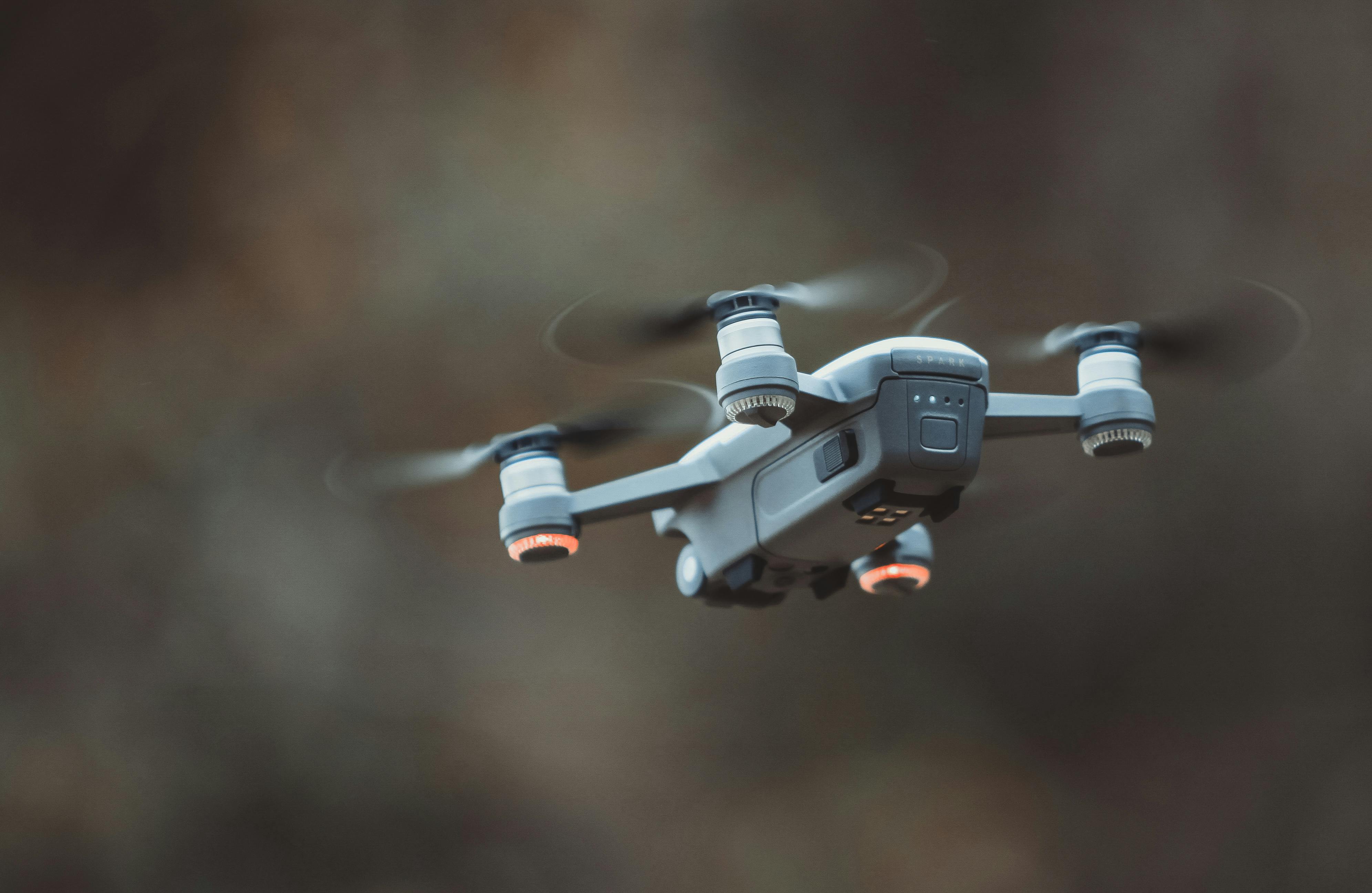 Small gray drone with spinning propellers flying against a blurred brown background.