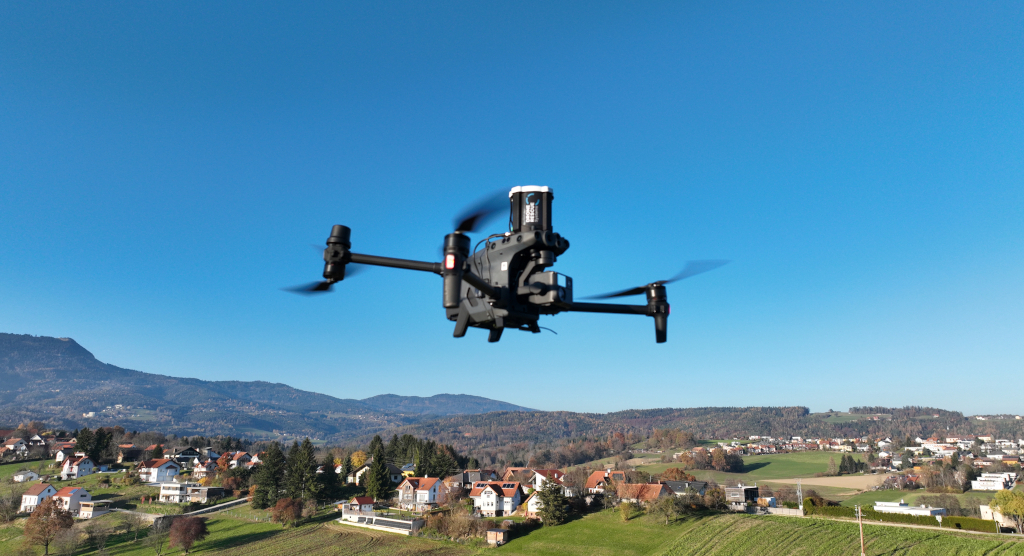 Black quadcopter drone flying over a rural landscape with houses and hills under a clear blue sky.