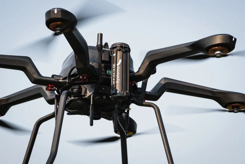 Close-up of a black multi-rotor drone flying with spinning propellers against a clear sky.