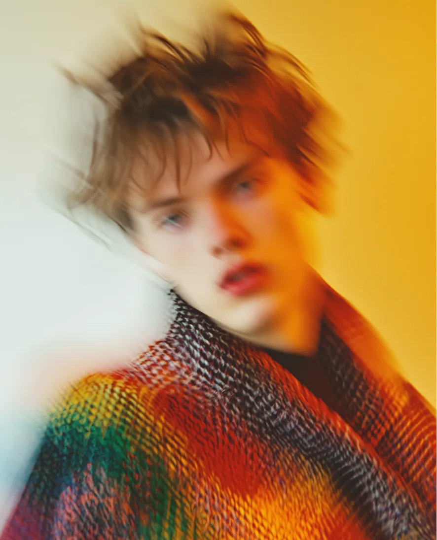 Blurred portrait of a person with tousled hair wearing a colorful patterned garment against an orange background.