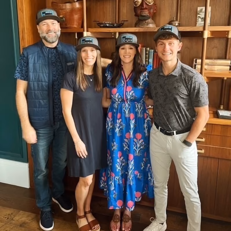 Four people standing indoors, smiling, wearing matching caps; two women and two men in casual attire.